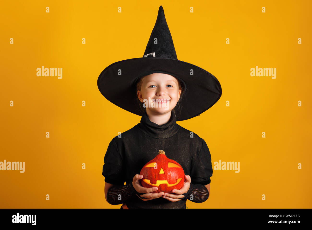Cute little girl dressed in a witch costume holds a pumpkin jack lantern on a yellow background. Halloween celebration. Stock Photo