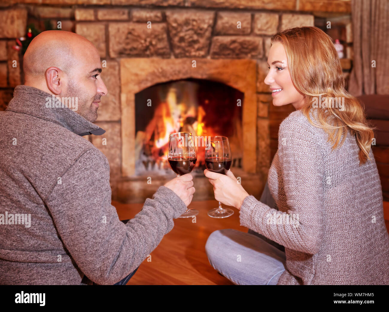Side view of happy cheerful couple sitting on the floor near fireplace, clinking glasses with tasty red wine, enjoying romantic date Stock Photo