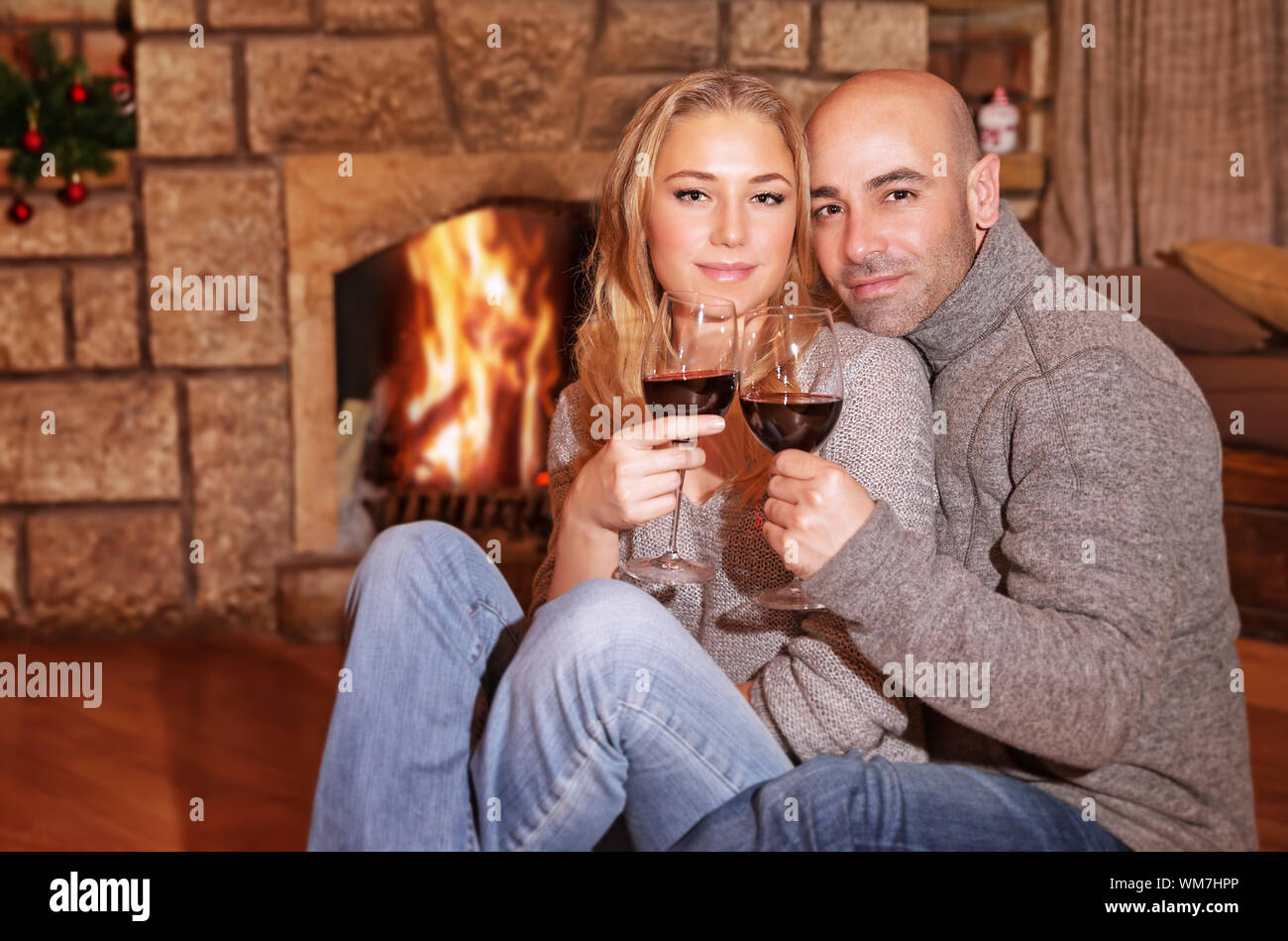 Cute couple on romantic date, beautiful woman with handsome man sitting near fireplace and drinking wine, celebrating Christmas holidays Stock Photo
