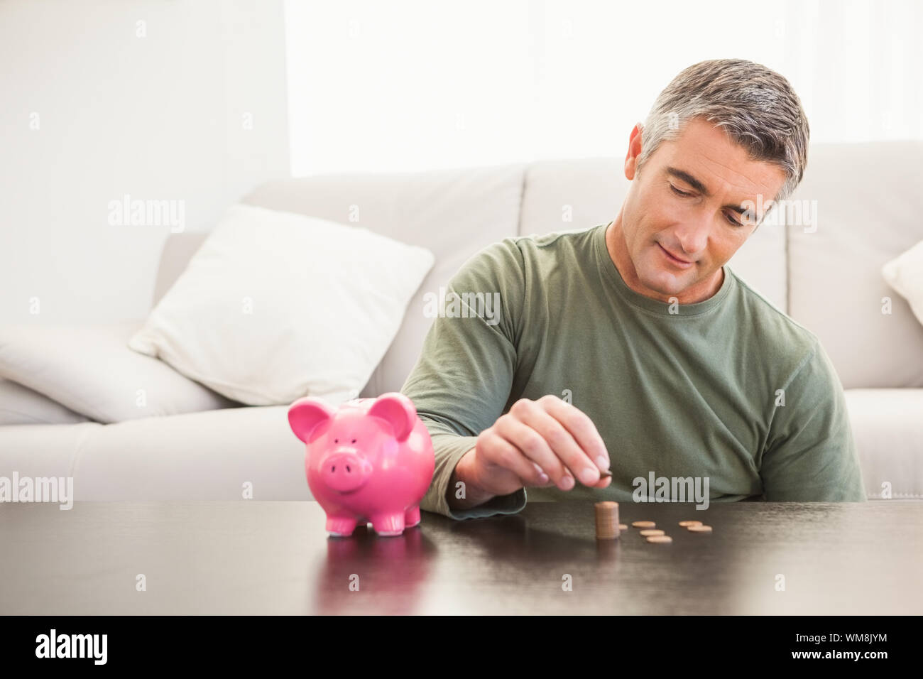 Smiling man putting some coins into a piggy bank at home in the living ...
