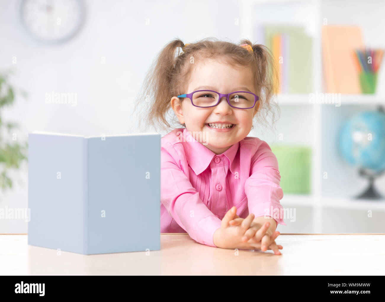 Happy kid girl in eyeglasses reading with standing book on her table Stock Photo