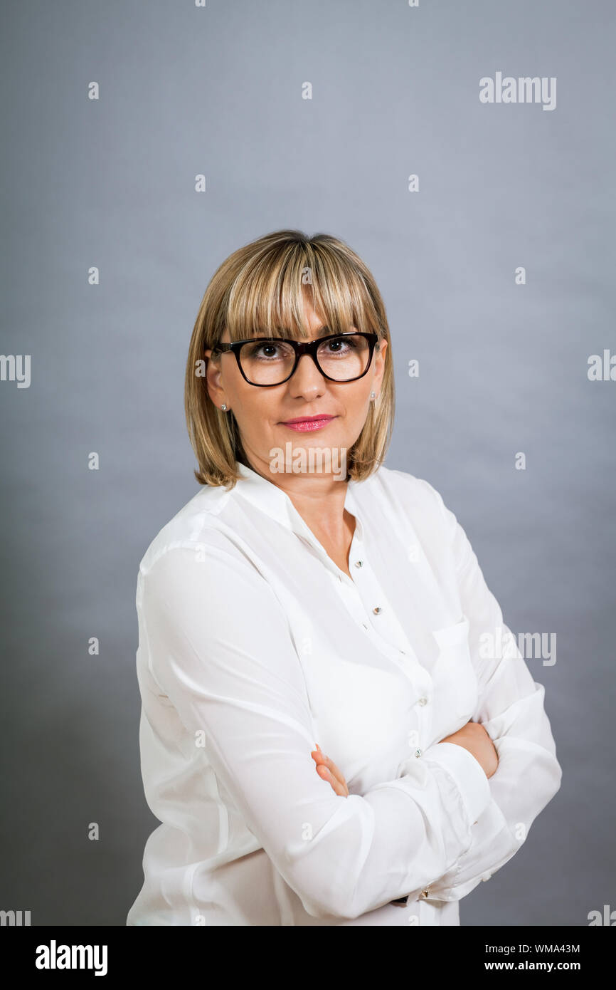 Scholarly attractive middle-aged blond woman in glasses standing looking at the camera with folded arms against a grey background Stock Photo