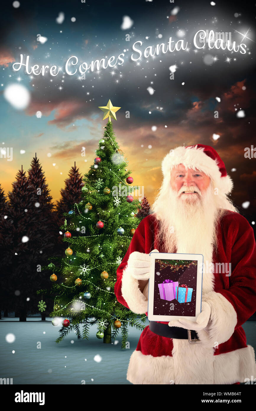 Santa presents a tablet PC against christmas tree in snowy landscape ...