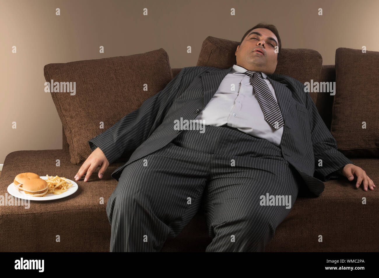 Obese man in formal clothes sleeping on sofa with plate of burgers and french fries by his side Stock Photo