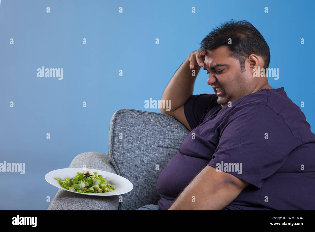 Side view of obese man sitting on sofa with hand on forehead showing dislike to green vegetable salad in plate Stock Photo
