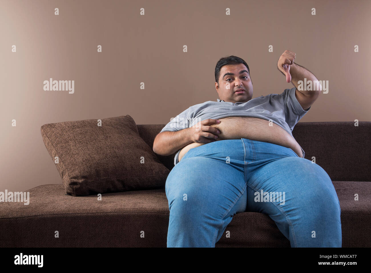 Unhappy obese man sitting on sofa holding his belly fat with one hand and showing thumbs down sign with other Stock Photo