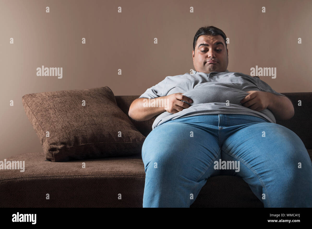 Obese man sitting on sofa holding his belly fat with both hands and looking  sad Stock Photo - Alamy