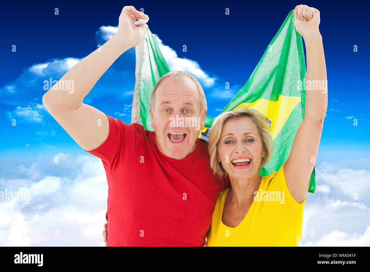 Happy older couple cheering at camera holding brazil flag against bright blue sky with clouds Stock Photo