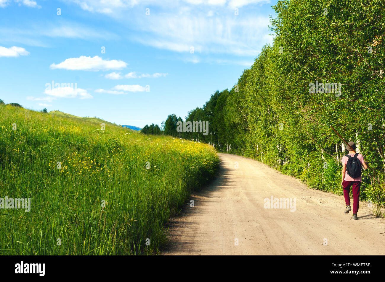 A Guy in a Pink T-Shirt, Red Pants, and Bucket Hat, with Black Backpack and  Long Pole on His Shoulder Walks along Highland Country Road. Sunny Spots o  Stock Photo - Alamy