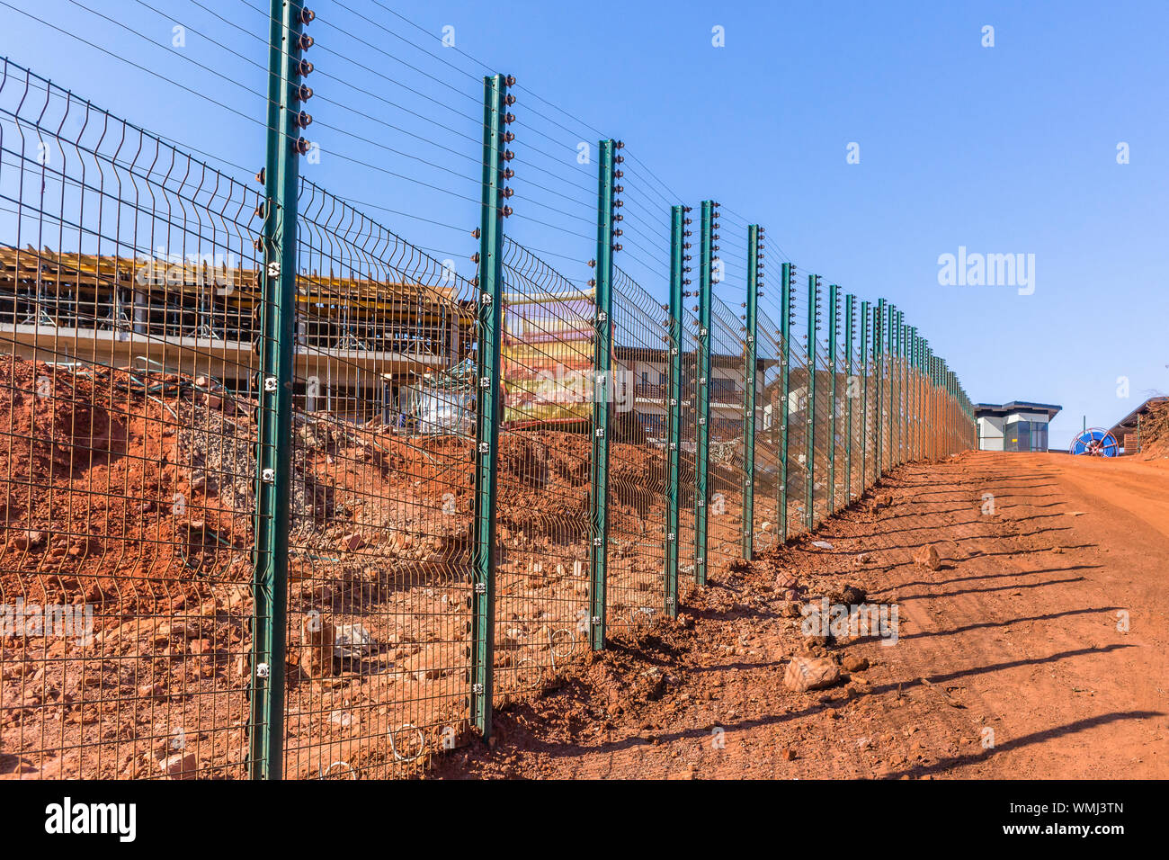 New Security electrified fence alongside dirt road with building construction of countryside living apartments in progress with blue sky Stock Photo