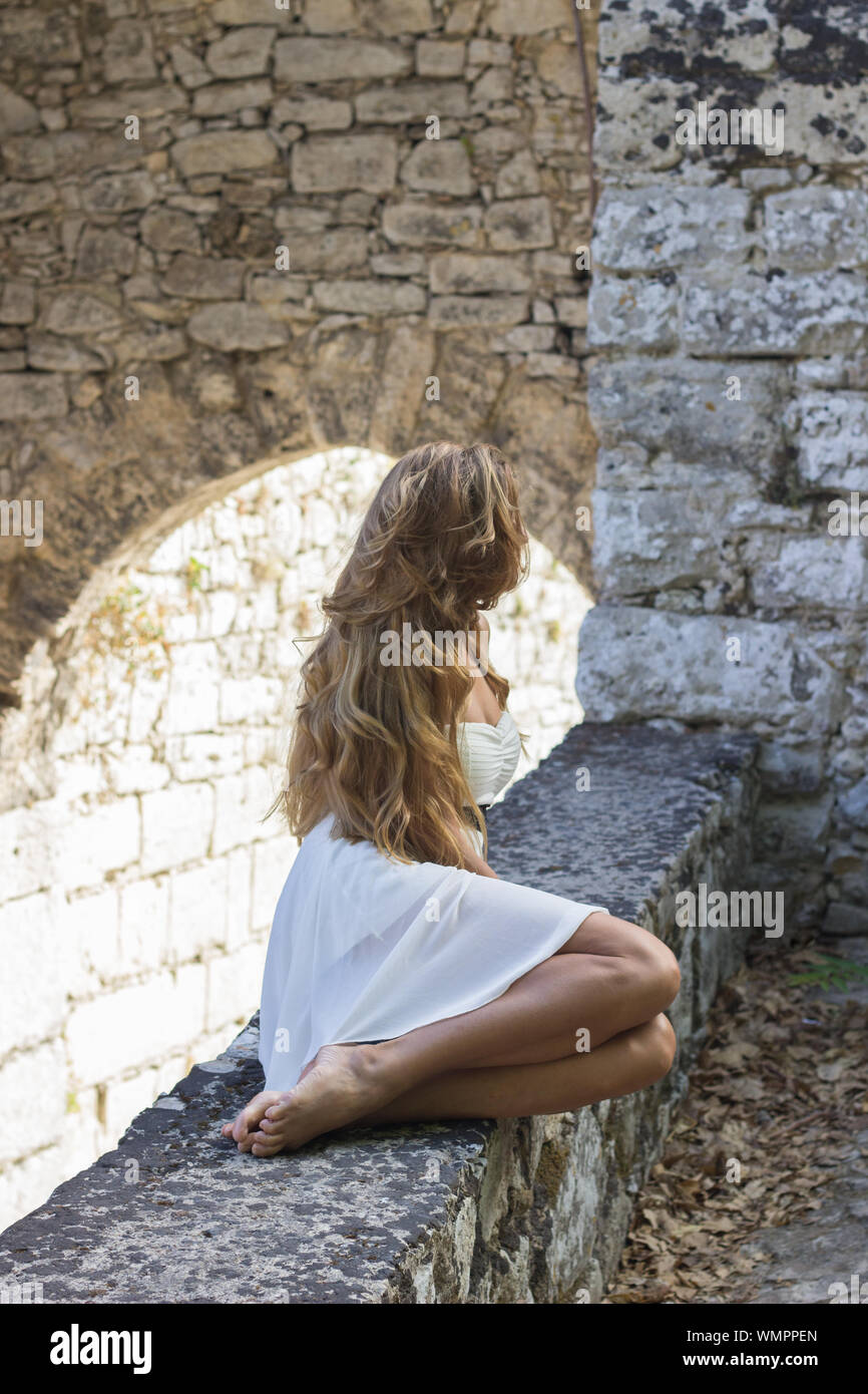 Young woman wearing white dress sitting on stone wall at ancient castle ard Stock Photo