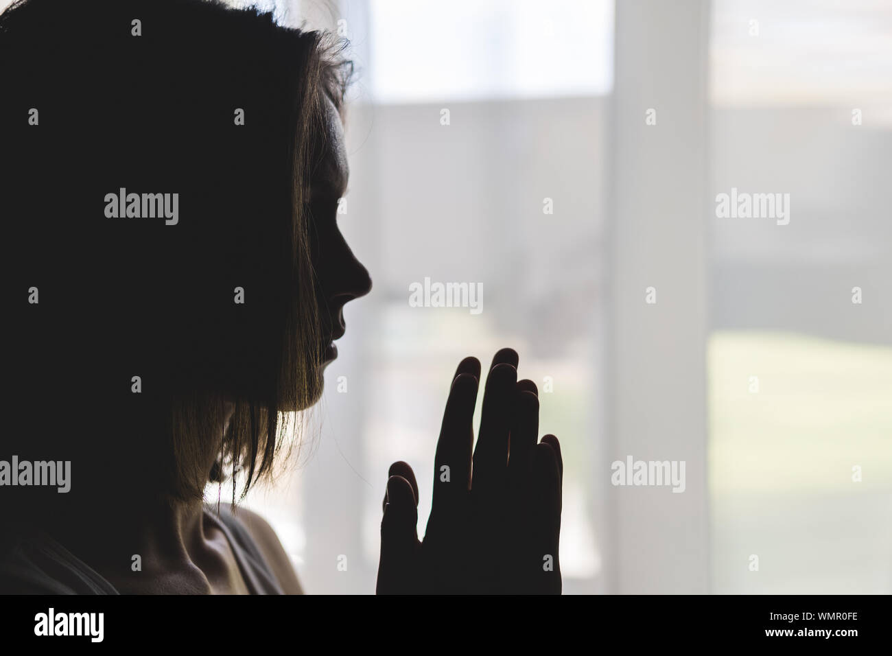 Silhouette of girl at window of clasped hand in gesture of prayer Stock Photo