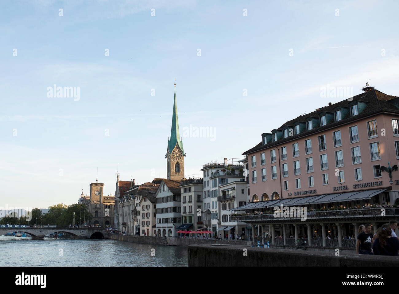 The spire of the Fraumünster Church at sunset, with shops and the Storchen Hotel on the Limmat River and Münsterbrücke bridge, in Zurich, Switzerland. Stock Photo