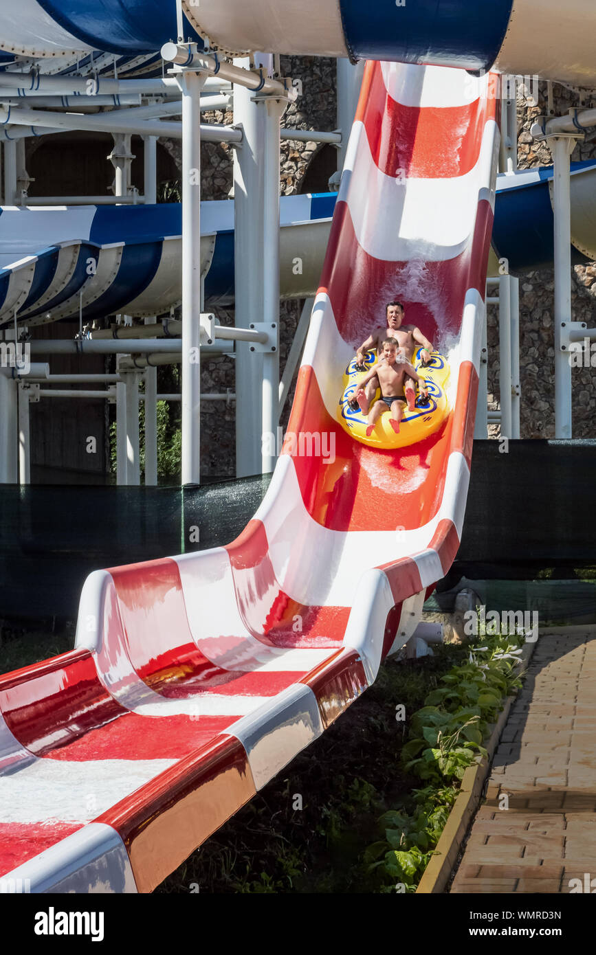 Family is riding down the water park red and white striped steep structure, sitting together on yellow inflatable ring. Father and son, adult and teen Stock Photo