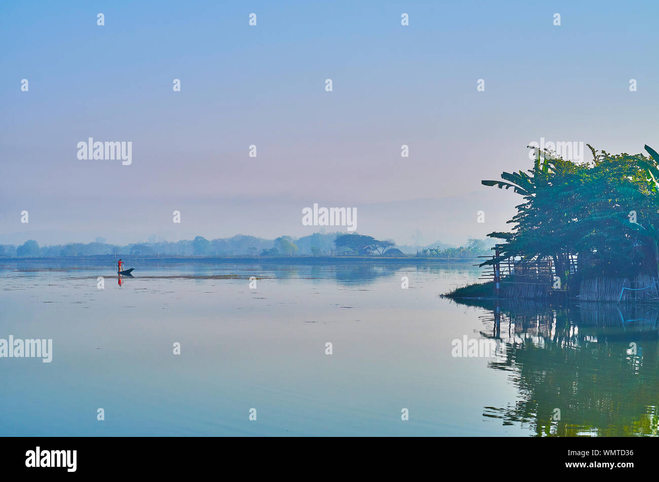 The clear surface of Tharzi pond reflects the hazy dawn sky, traditional Burmese fisherman in kayak is seen on background, Nyaungshwe, Myanmar. Stock Photo