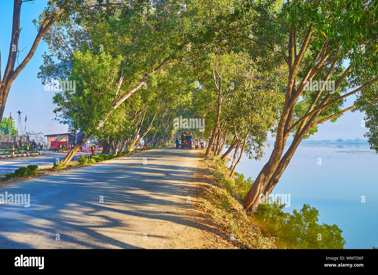 The shady road at Tharzi pond with lush thickets of trees, cars stop to make donations to workers of local Buddhist temple, Nyaungshwe, Myanmar Stock Photo