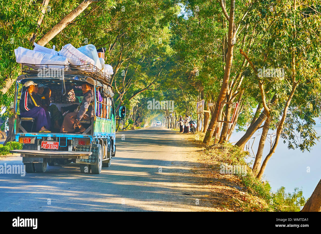 NYAUNGSHWE, MYANMAR - FEBRUARY 20, 2018: The road in scenic natural location at Tharzi pond with lush green trees on both sides and riding truck with Stock Photo
