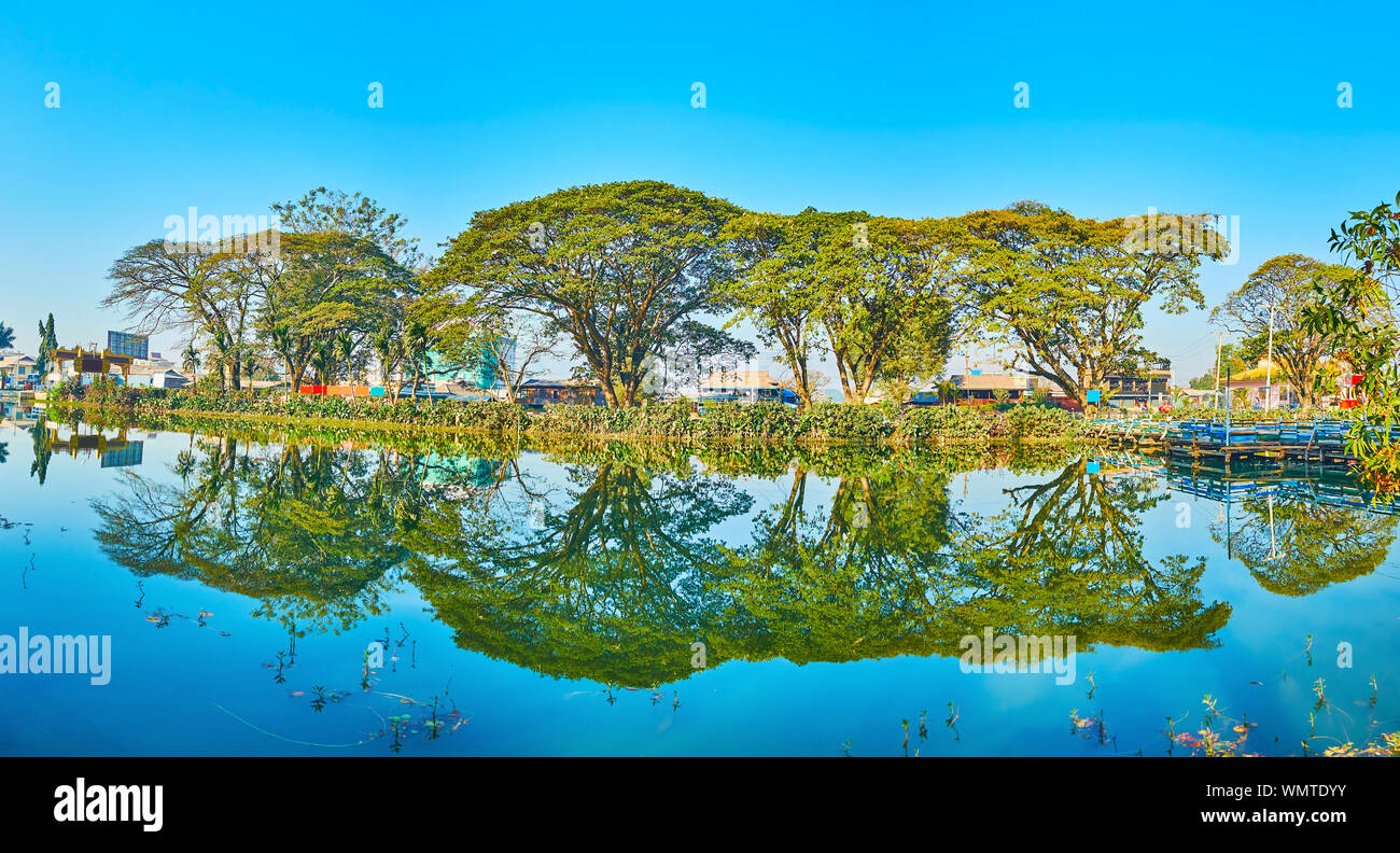 Panorama of Tharzi pond, its green embankment with line of old spread trees, providing the shade and reflected on blue surface, Nyaungshwe, Myanmar Stock Photo