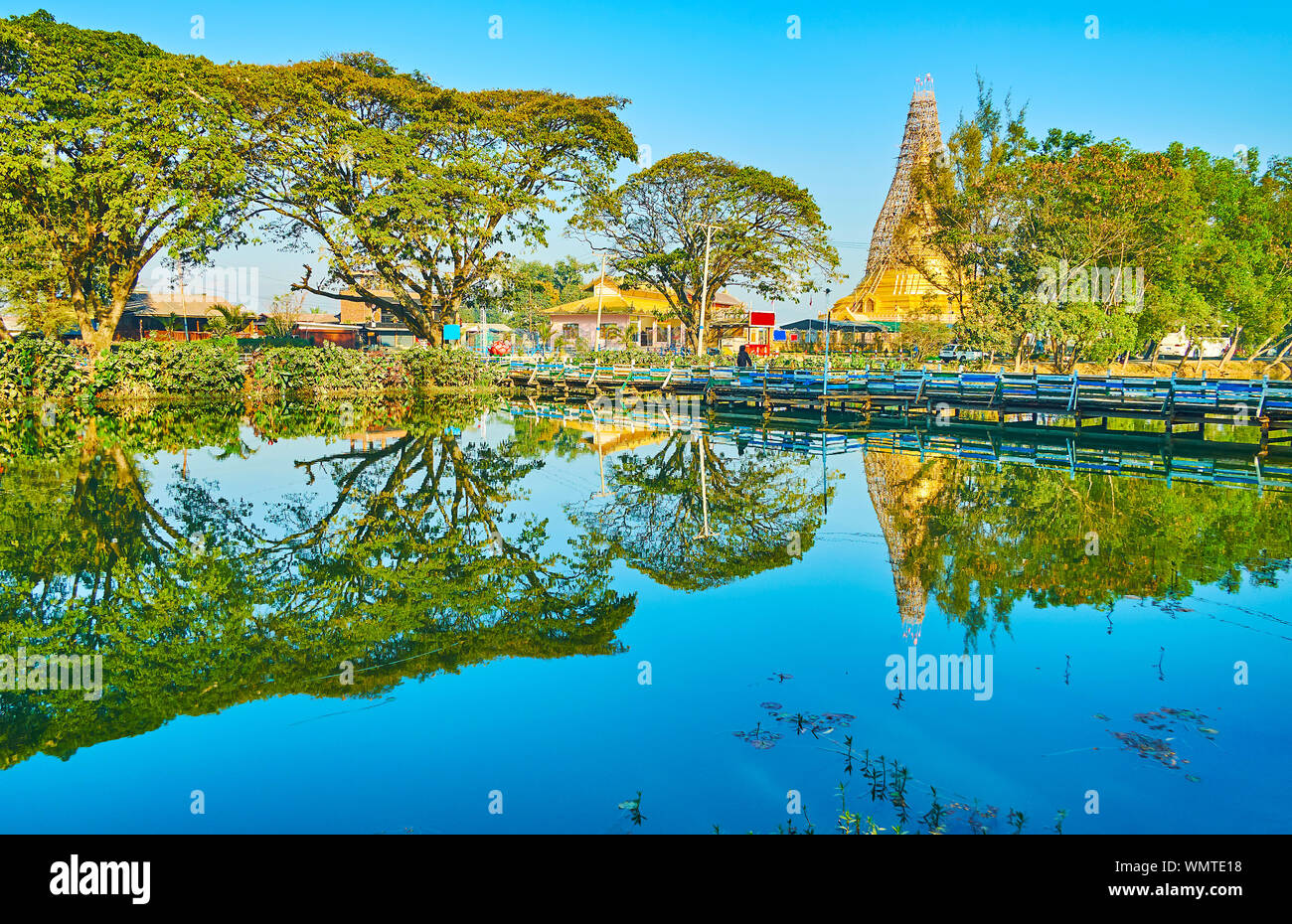 The pleasant walk by the Tharzi pond, surrounded by greenery, that hides golden pagoda of Buddhist Temple, Nyaungshwe, Myanmar Stock Photo