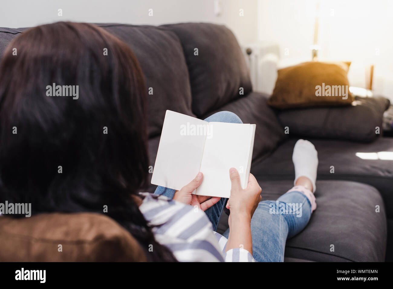 Young Latina girl reading a book lying on the sofa at home on a sunny day. Lifestyle. Stock Photo