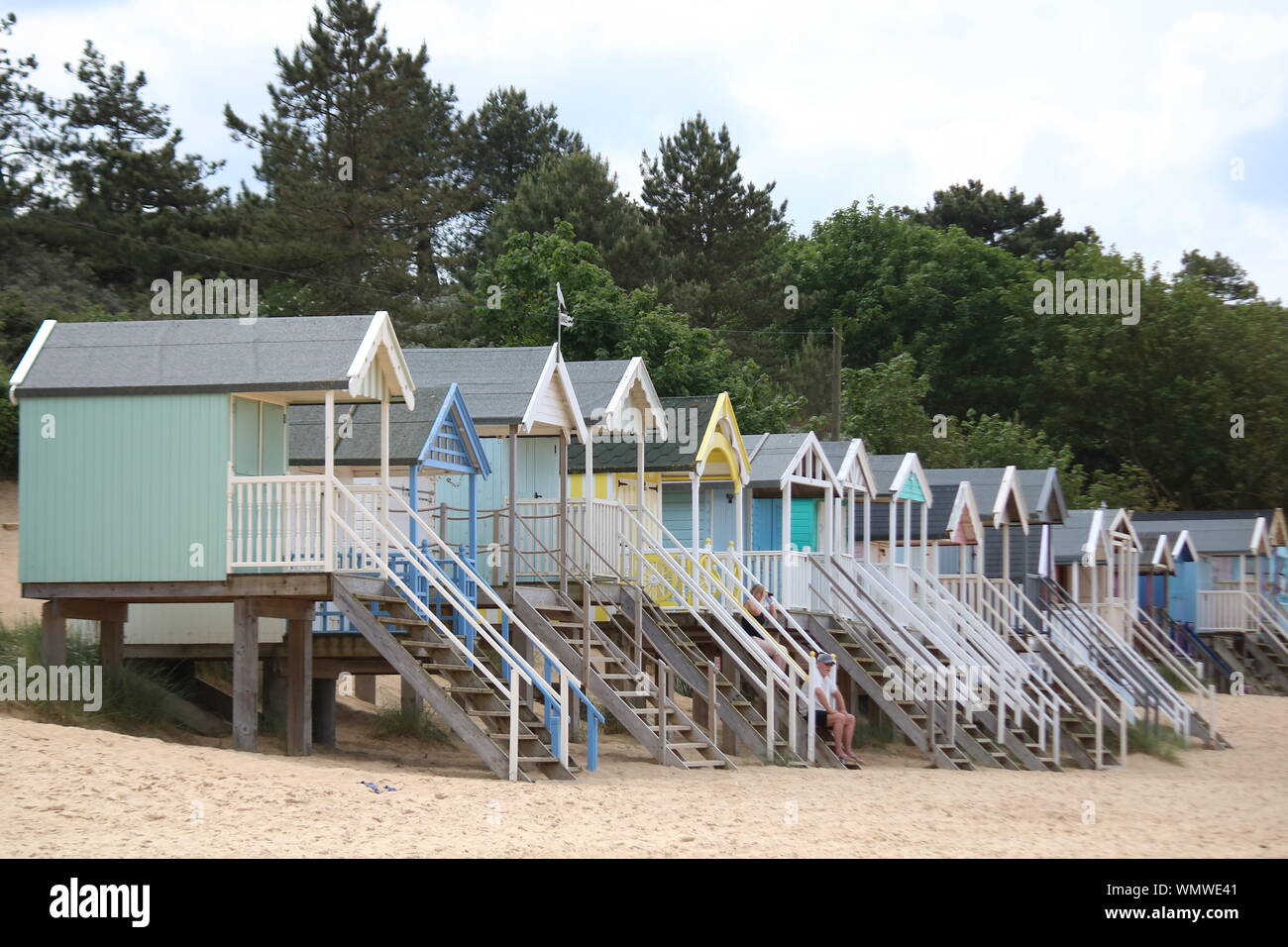 Row of traditional pastel coloured beach huts with two people on the steps of one of them on a beach at Wells-next-to the sea in Norfolk Stock Photo