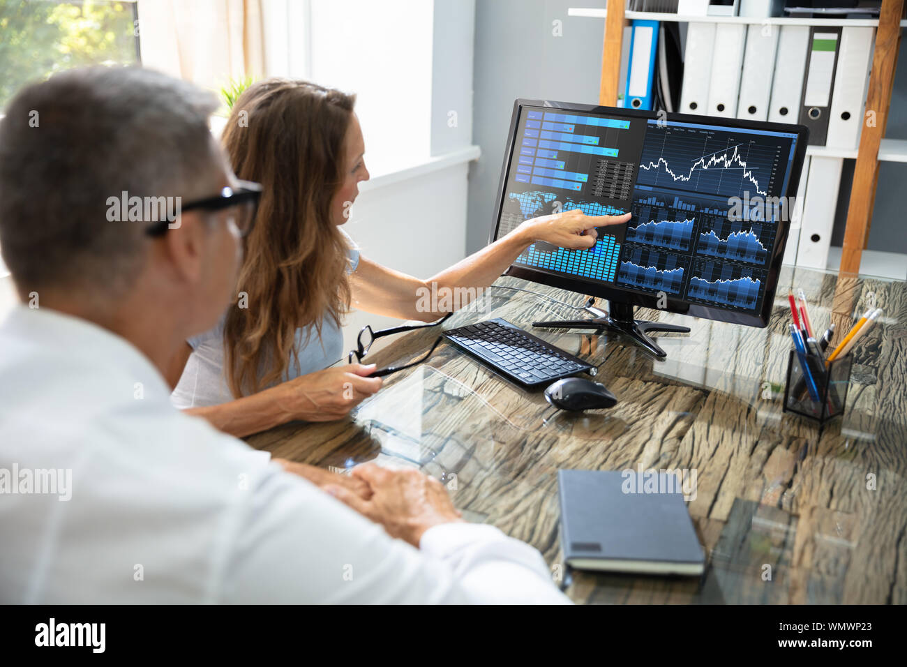 Side View Of Two Stock Market Brokers Discussing Graphs On Computer At Workplace Stock Photo