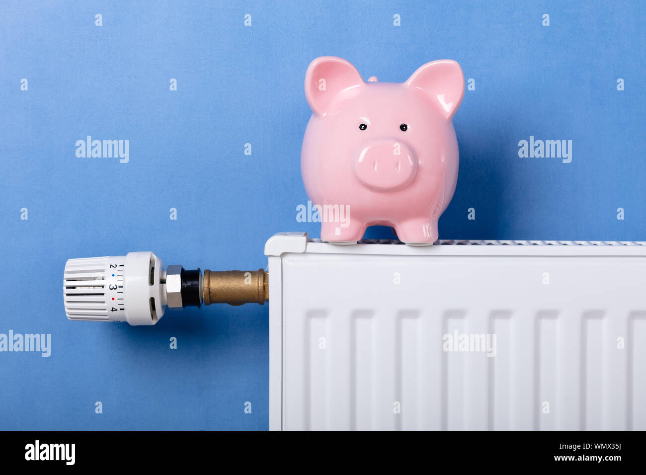 Piggy Bank On Heating Radiator With Temperature Regulator Against Blue Background Stock Photo