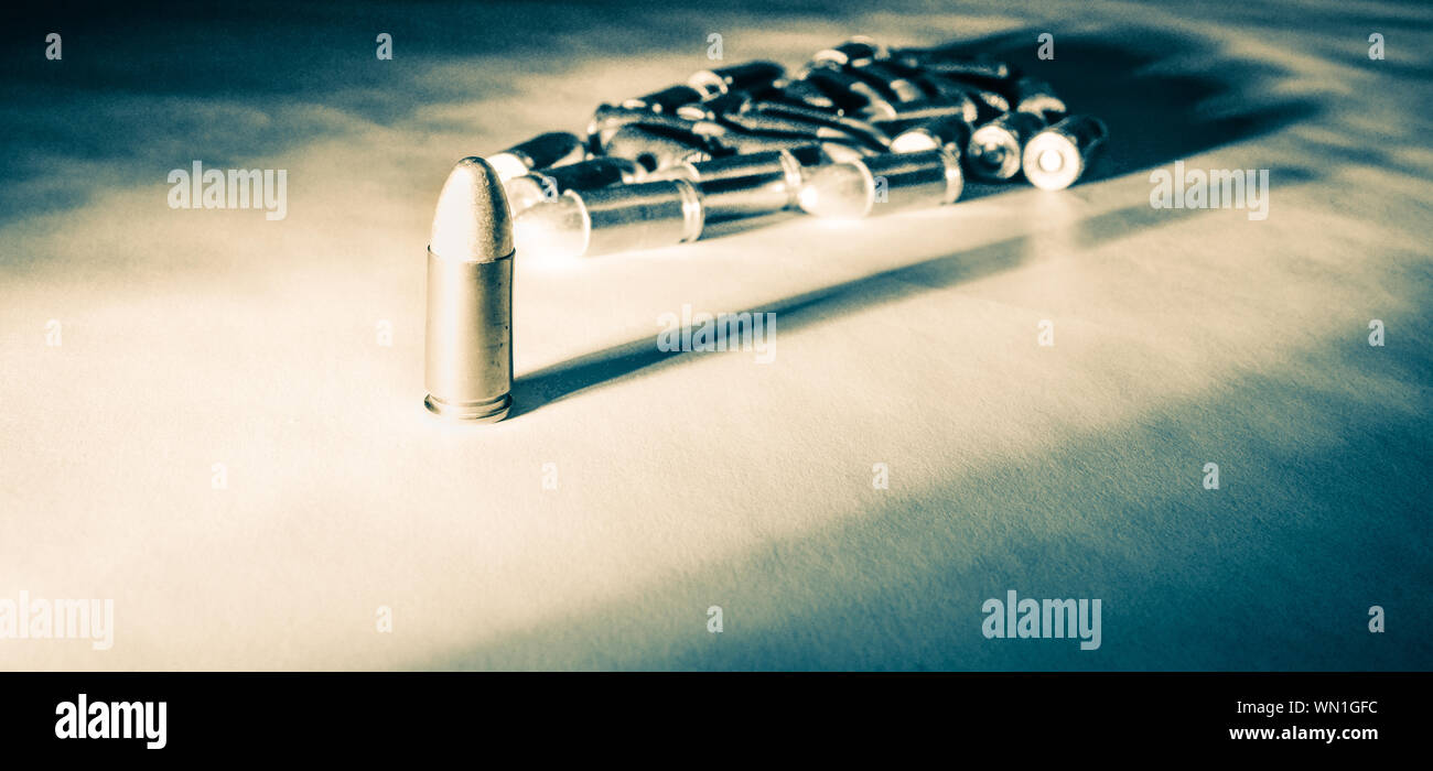 A single 9mm bullet stands alone with strong side light creating long shadows with a pile of bullets in background in greenish tone black and white Stock Photo