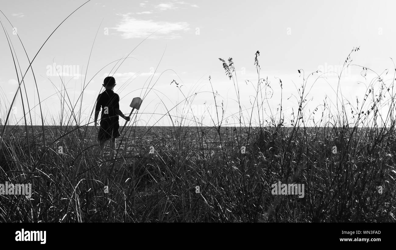 Grass Against Silhouette Boy With Shovel Standing At Beach Stock Photo