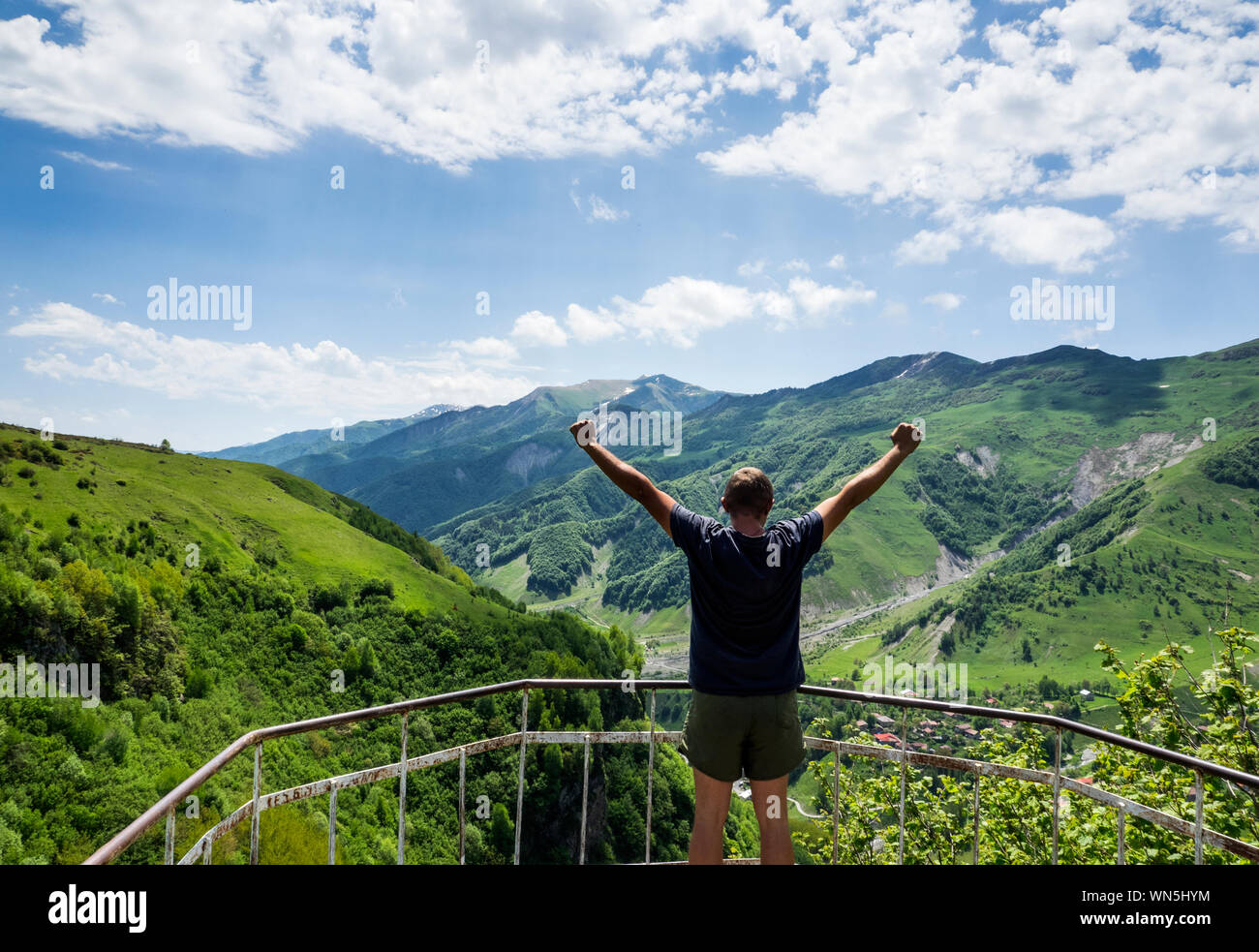 Man With Arms Raised Standing At Observation Point Against Mountain And Cloudy Sky Stock Photo