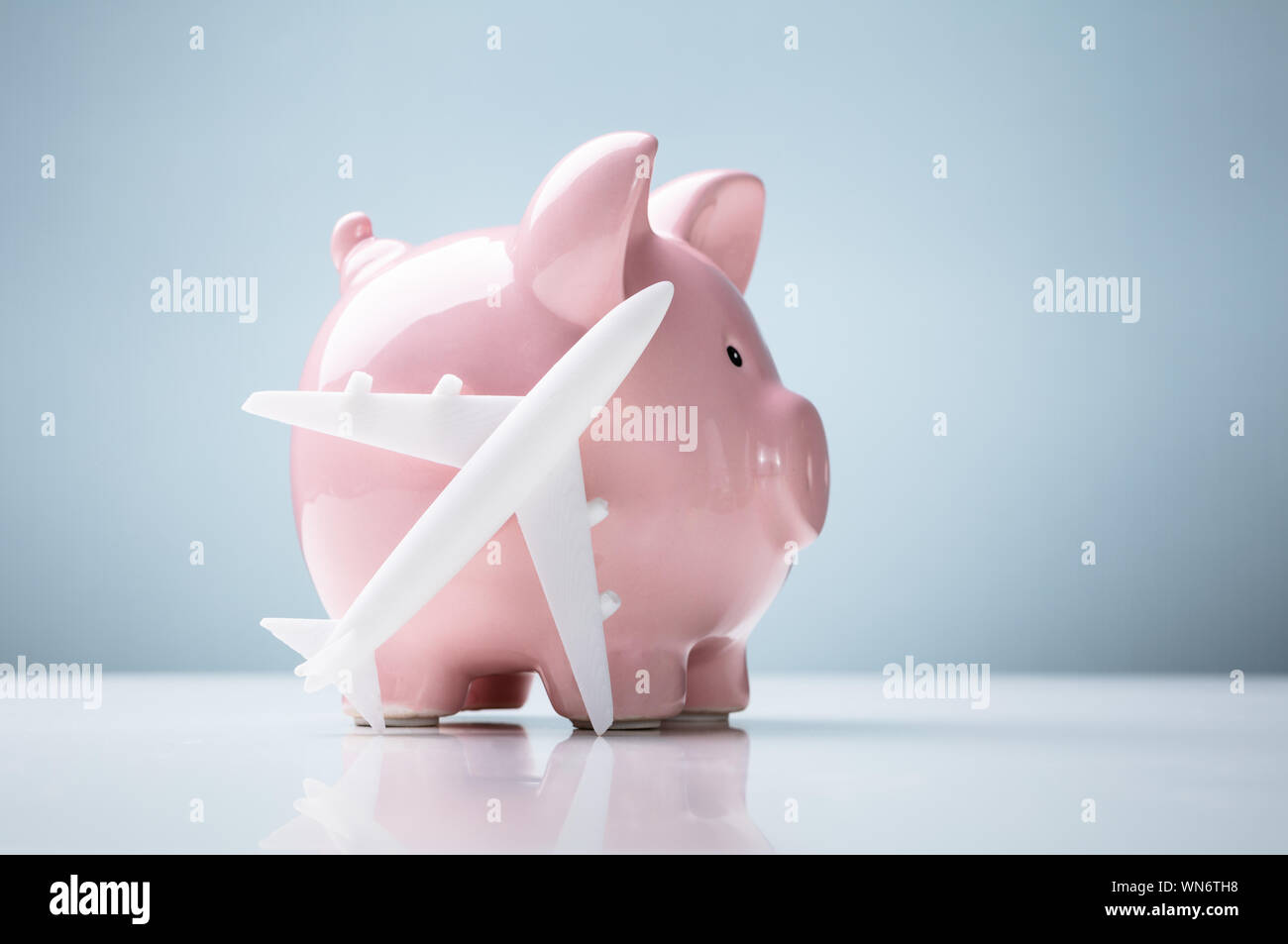 Close-up Of Piggy Bank With Airplane Over White Desk Against Blue Background Stock Photo