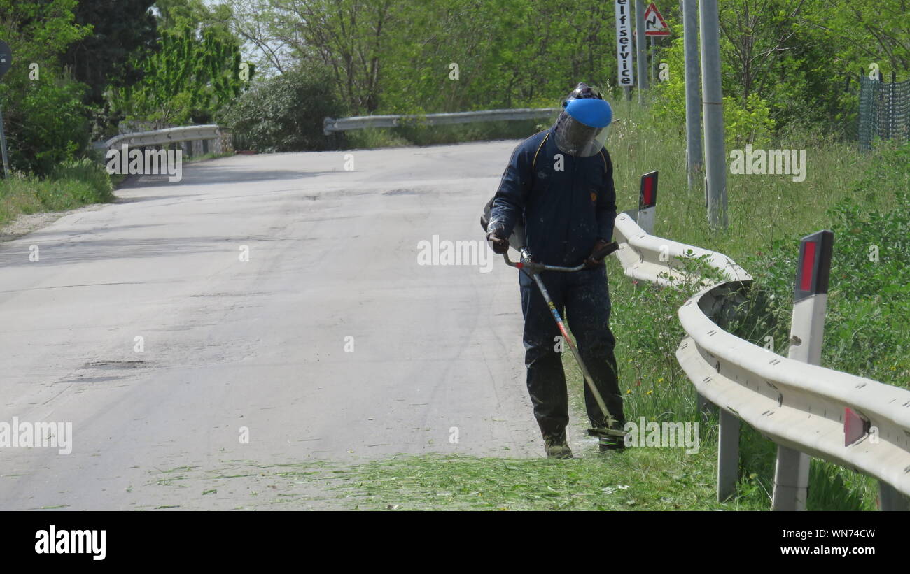 Worker Cutting Grass With Brush Cutter By Road Stock Photo