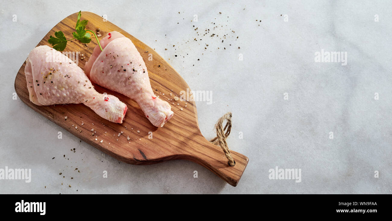 Two raw chicken legs or drumsticks with seasoning of chili, ground peppercorns and fresh herbs on a wooden board on marble with copy space, panorama b Stock Photo