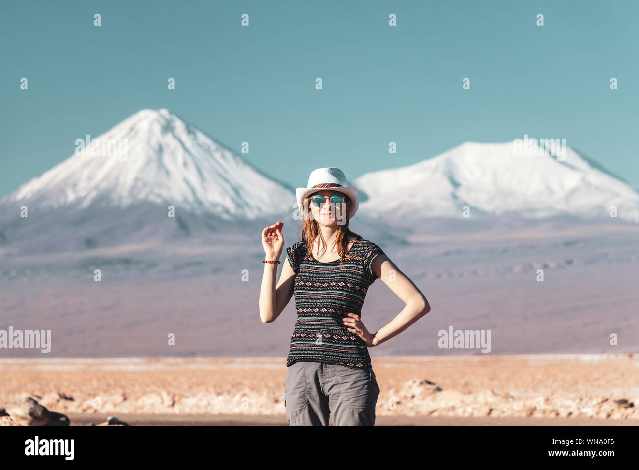 Young casual blond woman travel alone, caucasian girl in hat and sunglasses looking at camera with snow covered mountains on the background. Stunning Stock Photo