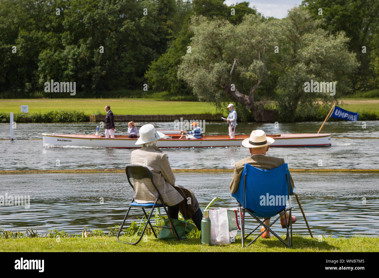 A senior couple sit on picnic chairs to watch the rowing at Henley Womens Regatta as the Umpire's Launch passes by. Stock Photo