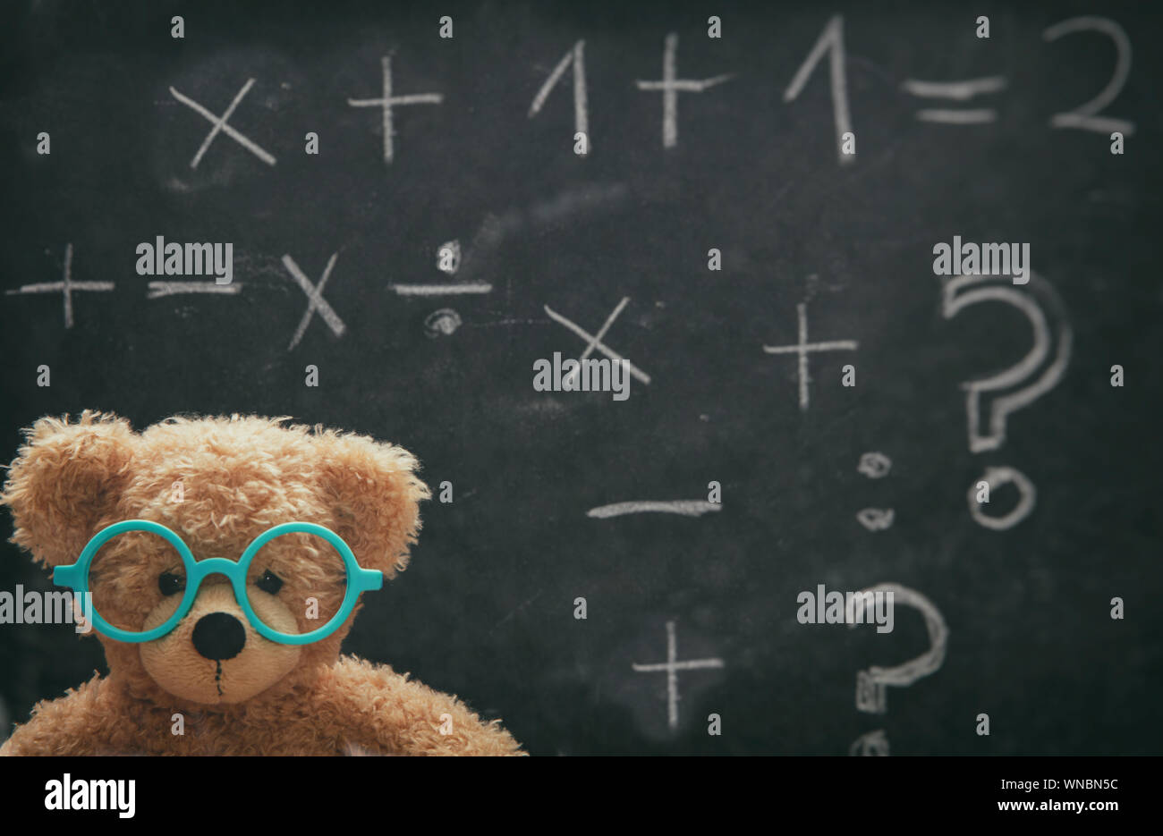 Maths concept. Smart kid in school class, cute teddy wearing blue eyeglasses and math symbols on blackboard Stock Photo