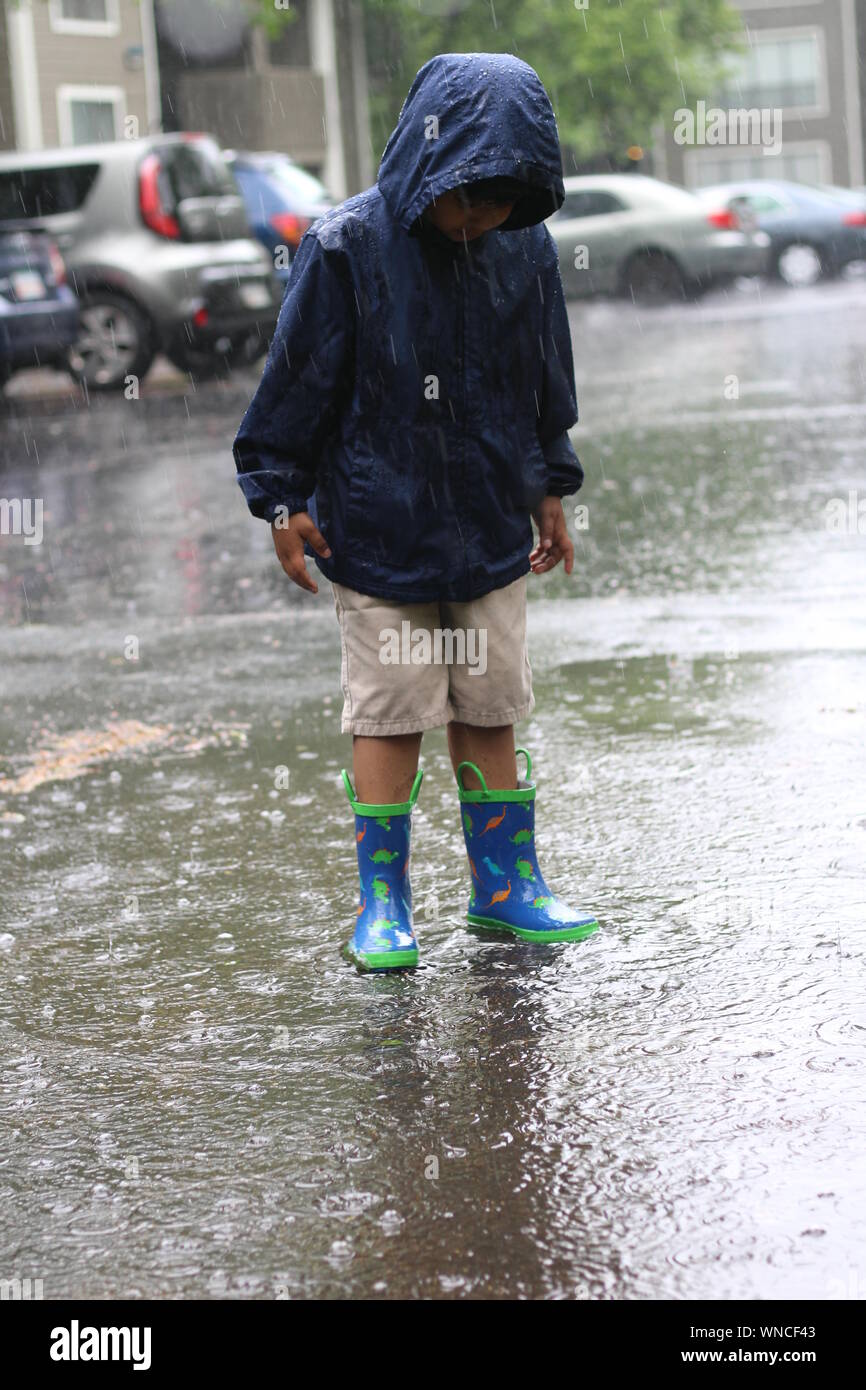 Boy Wearing Raincoat Standing On City Street During Rainy Season Stock