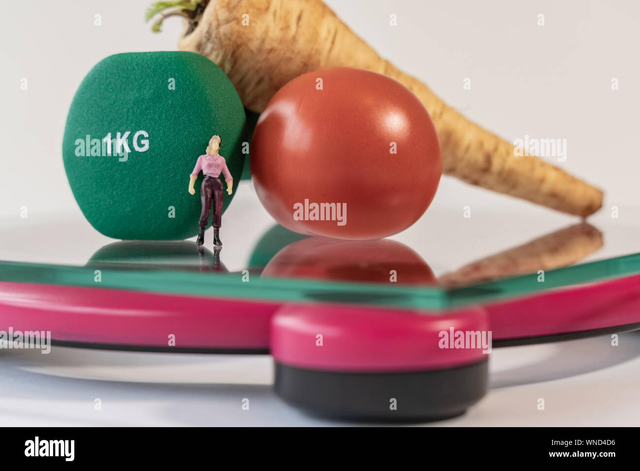 Miniature woman figure standing on the digital electronic bathroom scale for weight of human body. Fresh vegetables and green dumbbells at shallow dep Stock Photo