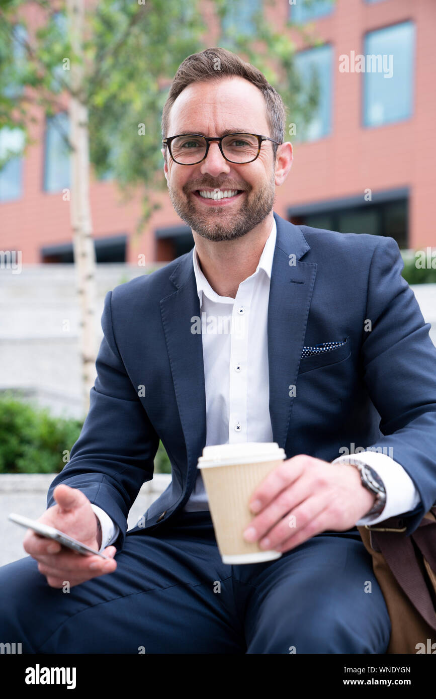 Portrait Of Mature Businessman With Mobile Sitting At Outside Office Building Stock Photo