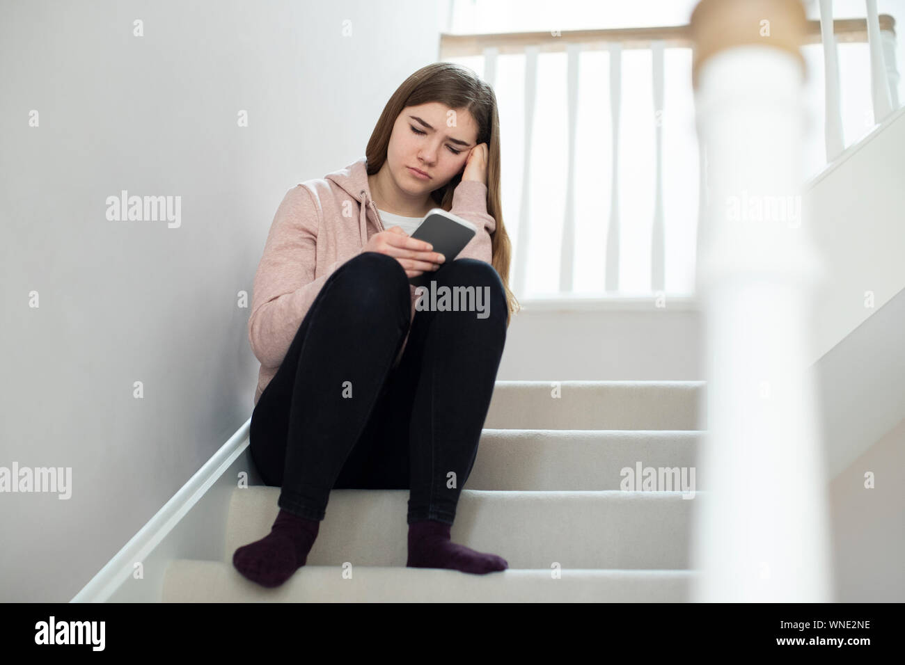 Unhappy Teenage Girl Being Bullied By Text Message Sitting On Stairs At Home Stock Photo