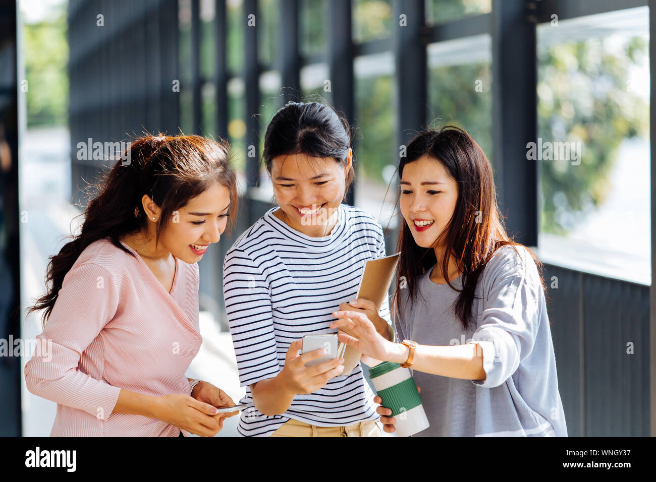 Young Asian business women talking and showing phone screen in office building in casual wear. Three girls having discussion outdoors Stock Photo