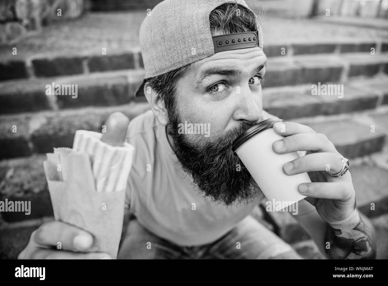 Junk food. Carefree hipster eat junk food while sit stairs. Snack for good mood. Guy eating hot dog. Street food concept. Man bearded eat tasty sausage and drink paper cup. Urban lifestyle nutrition. Stock Photo