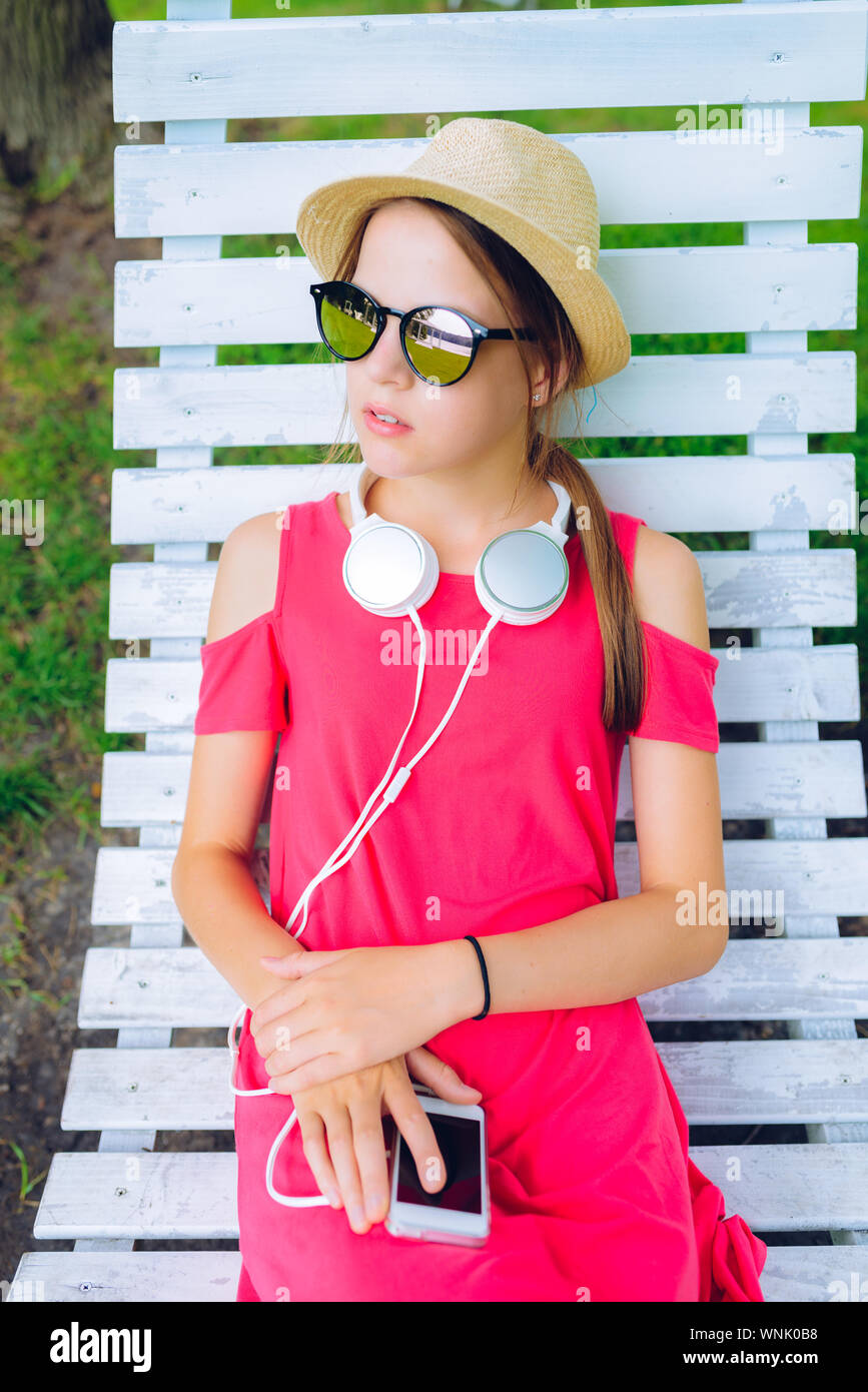 young girl resting on a hanging wooden chair in the garden - with headphones around her neck and a smartphone in hands Stock Photo