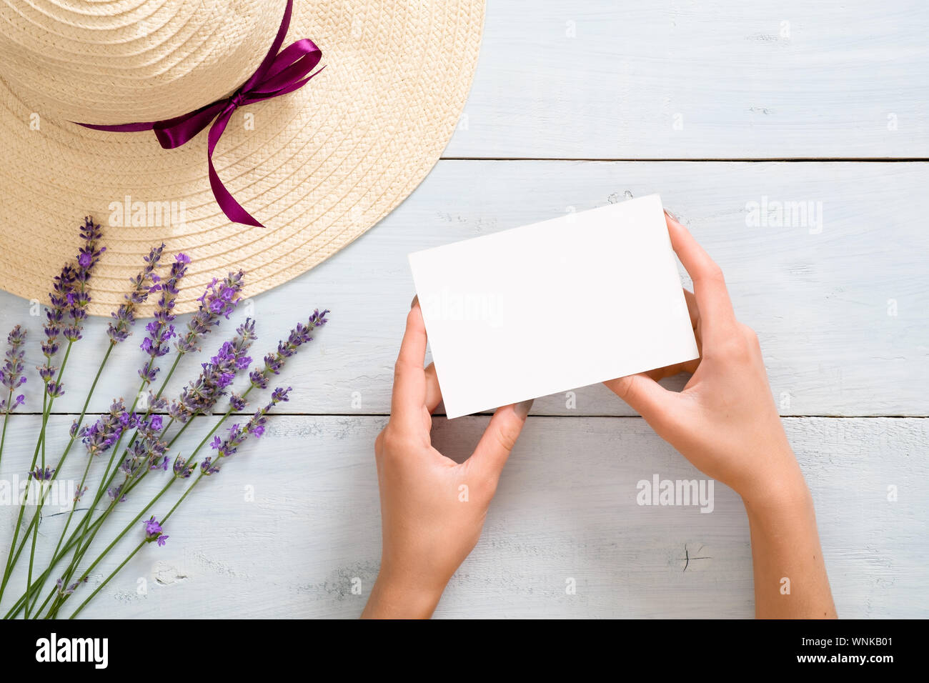 Hands of woman holding blank paper card mockup over rustic blue wooden background with straw hat and lavender flowers. Flat lay, top view, overhead. S Stock Photo