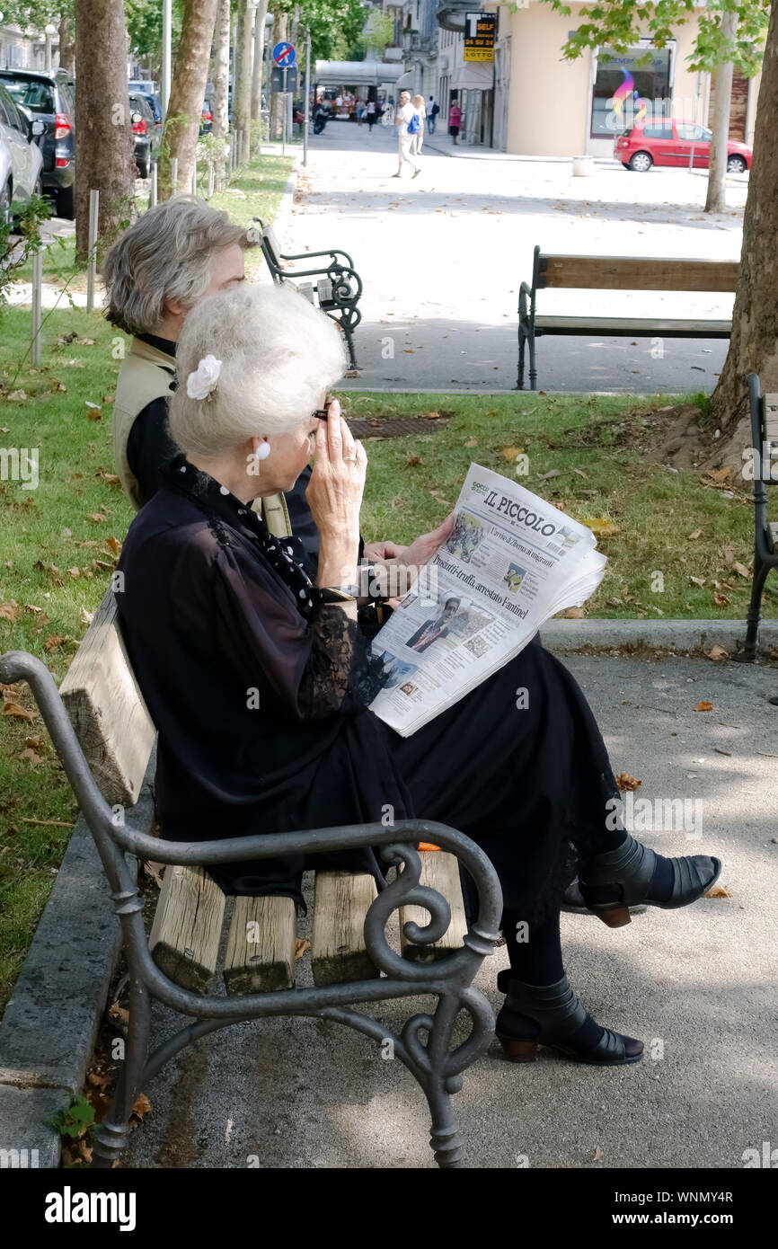 Elegant pretty elderly woman wearing old fashioned dress, sitting on a bench in a city public garden, reading the newspaper. 70, 80, 90 years old lady Stock Photo