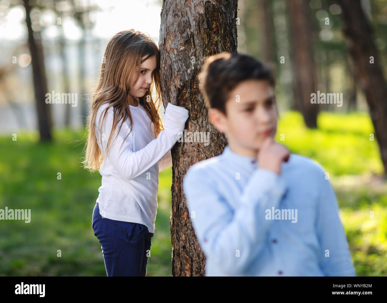 Boy and girl playing hide and seek in the park. Girl watching on boyfriend. Girl hiding behind the tree and lurk on boy. Stock Photo