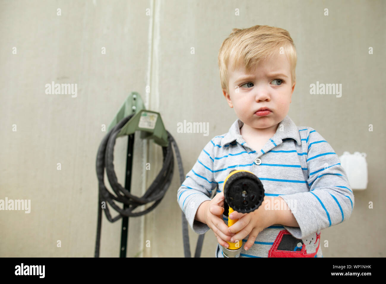 Upset toddler boy looks sideways while pouting and holding garden hose Stock Photo