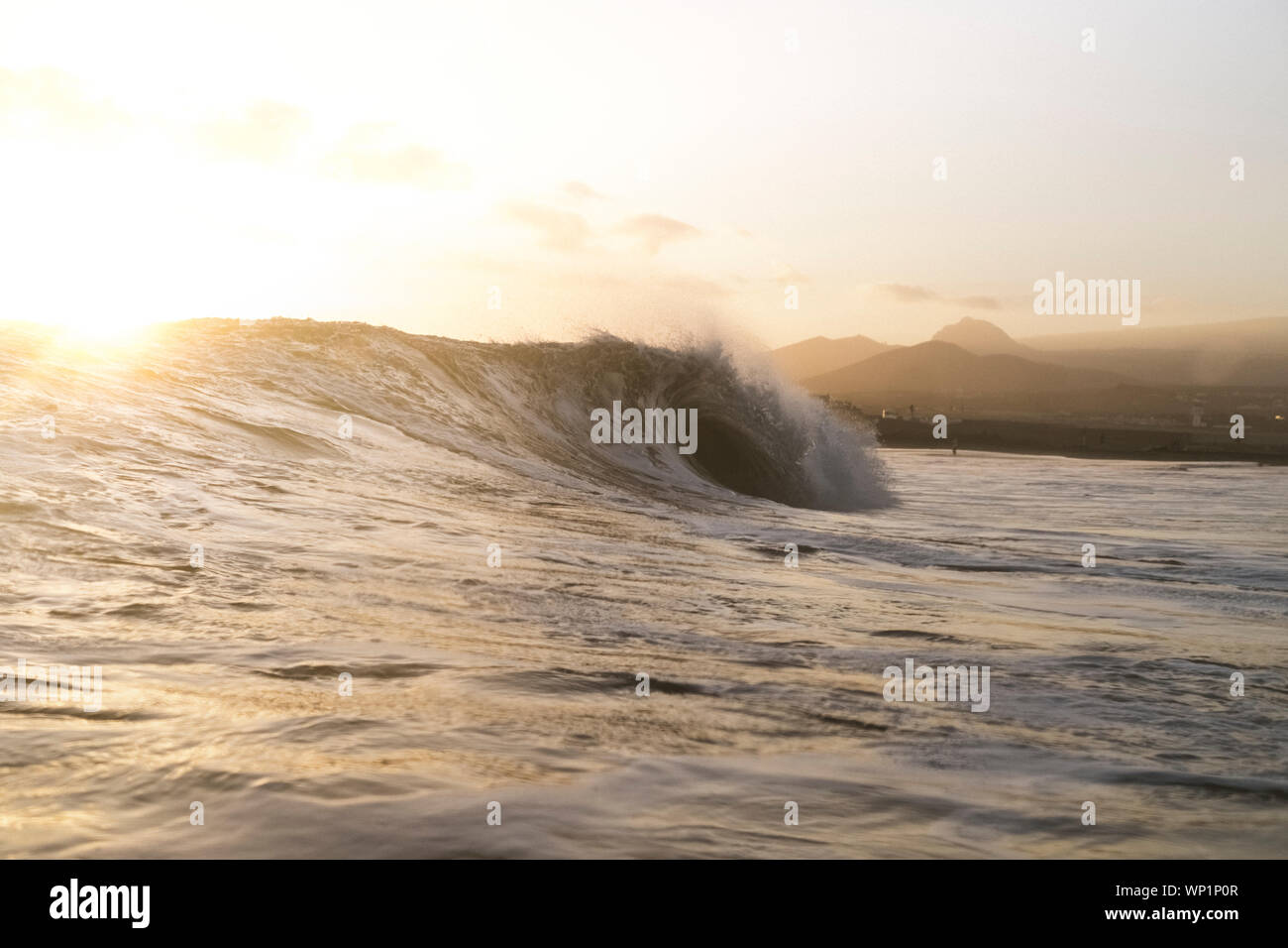 Backlit scene of wave breaking at sunset with shoreline in background Stock Photo