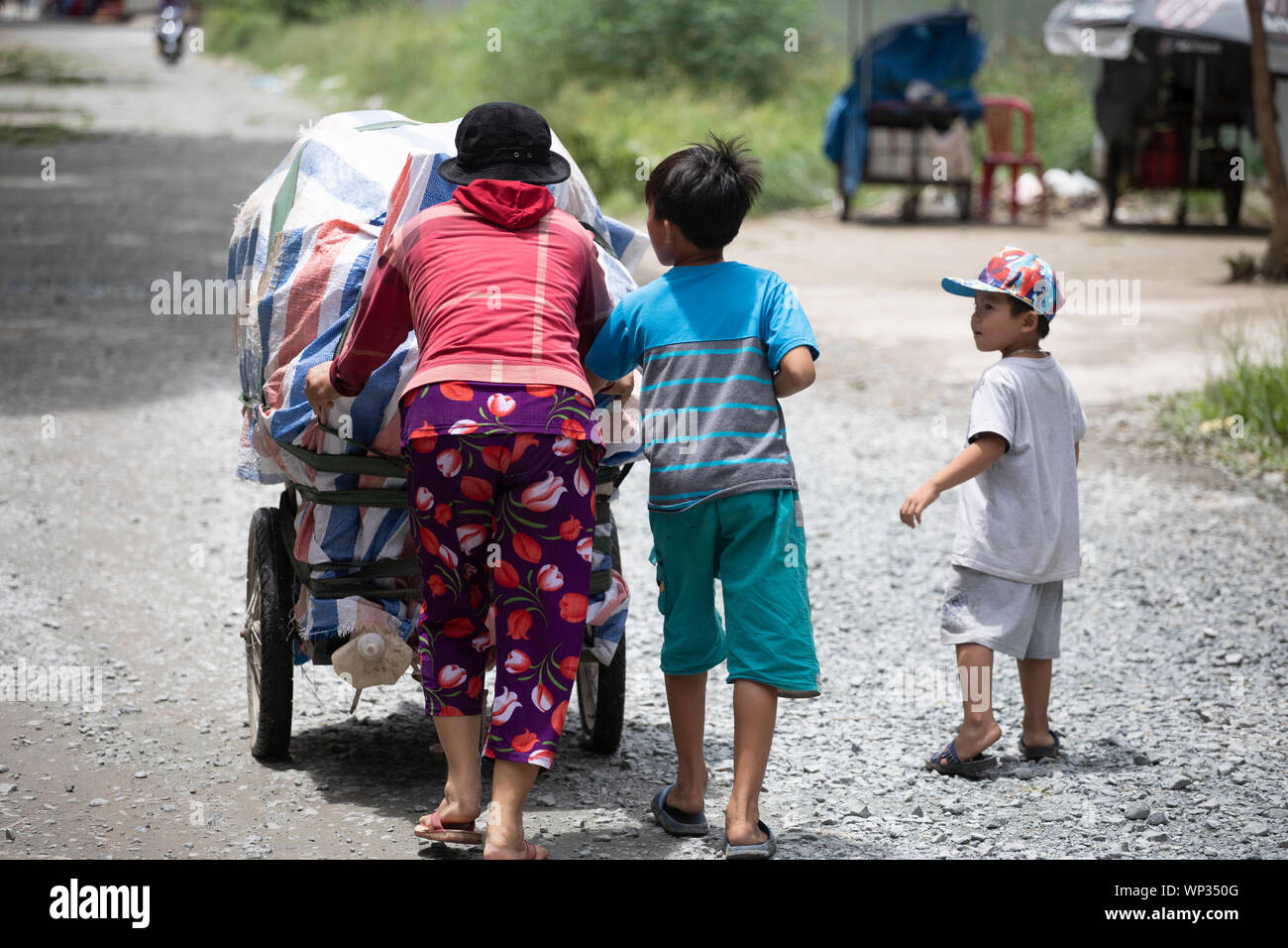 A mother and her two young sons push their packed up street food cart along a dirt road in district 7, Ho Chi Minh City after a busy day's cooking Stock Photo