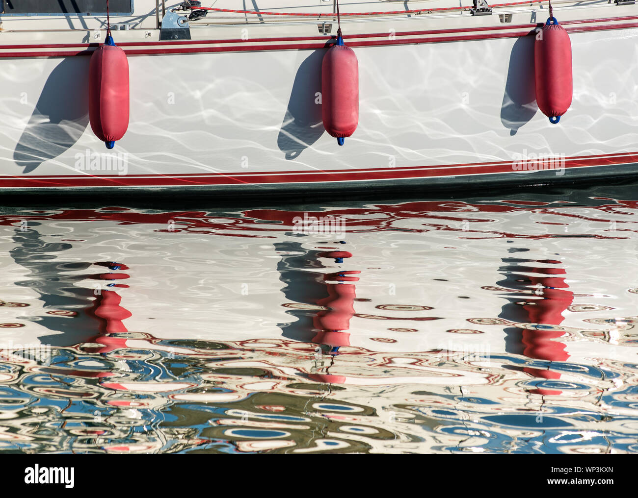 Three red fenders hanging over the hull of a boat reflecting in the sparkling sunlit water of